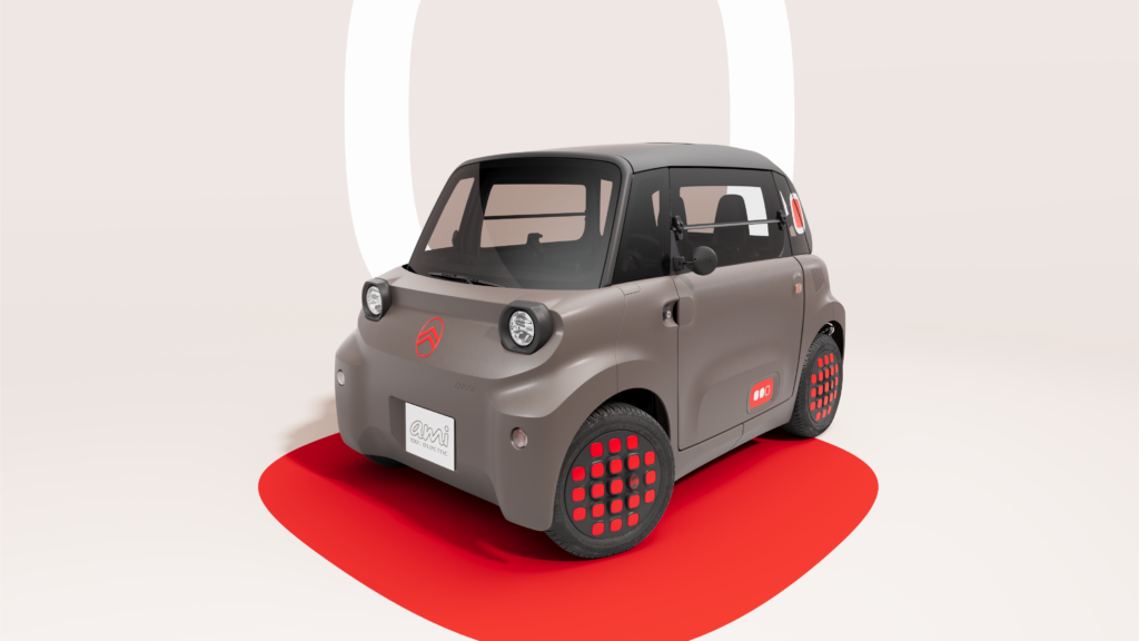 Auf diesem Bild ist ein kleines, graues Citroën Ami Elektroauto mit markanten roten Akzenten an den Radkappen und der Karosserie zu sehen, platziert auf einer roten, abgerundeten Fläche vor einem hellen Hintergrund.