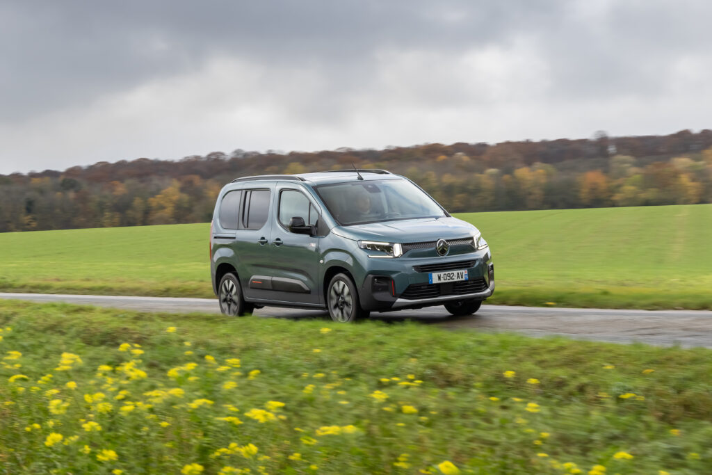 Grüner Citroën ë-Berlingo fährt auf einer Landstraße durch eine ländliche Landschaft.