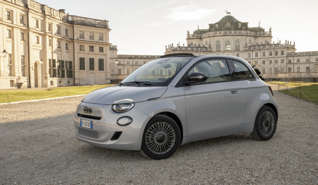 Weißer Fiat 500 Hybrid vor einem historischen Schlossgebäude mit weitläufigem Vorplatz.