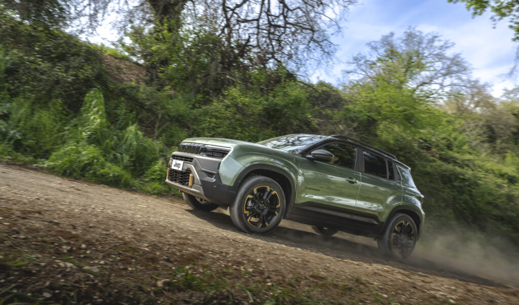 Grüner Jeep Avenger 4xe fährt über einen unbefestigten Waldweg und wirbelt Staub auf.