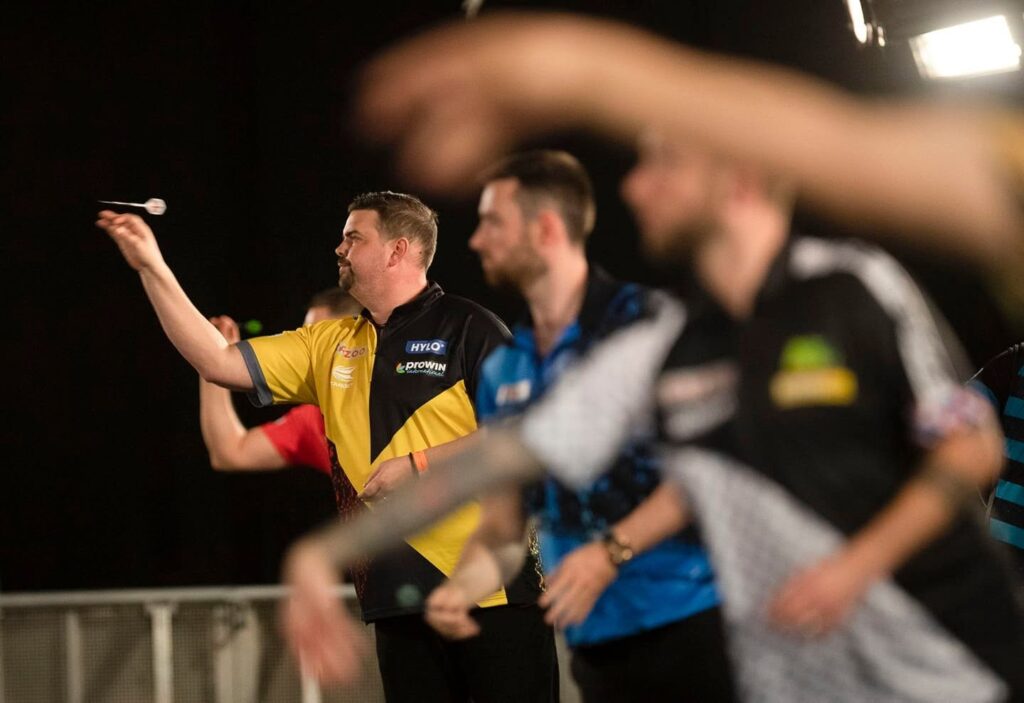 Ein Dartspieler im gelb-schwarzen Trikot wirft konzentriert einen Pfeil, während weitere Spieler im Vordergrund unscharf zu sehen sind.