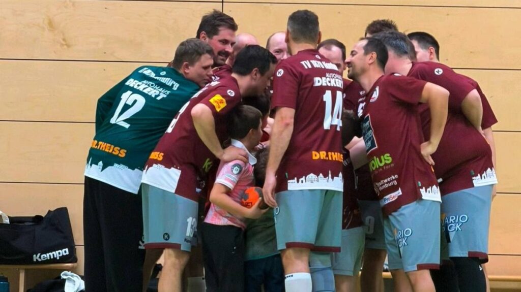 Handballspieler in dunkelroten Trikots stehen vor einem Spiel eng im Mannschaftskreis zusammen. Zwei Kinder befinden sich inmitten der Gruppe.