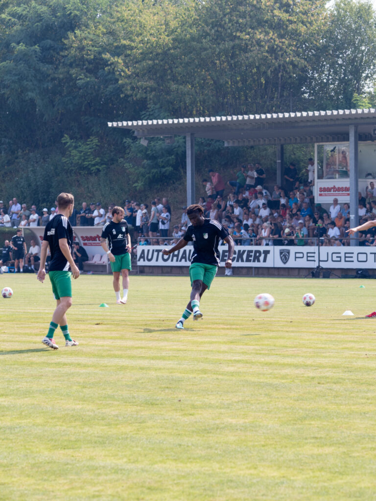 Fußballspieler in schwarzen Trikots und grünen Hosen wärmen sich auf dem Rasen auf. Im Hintergrund verfolgen zahlreiche Zuschauer das Training von einer überdachten Tribüne.