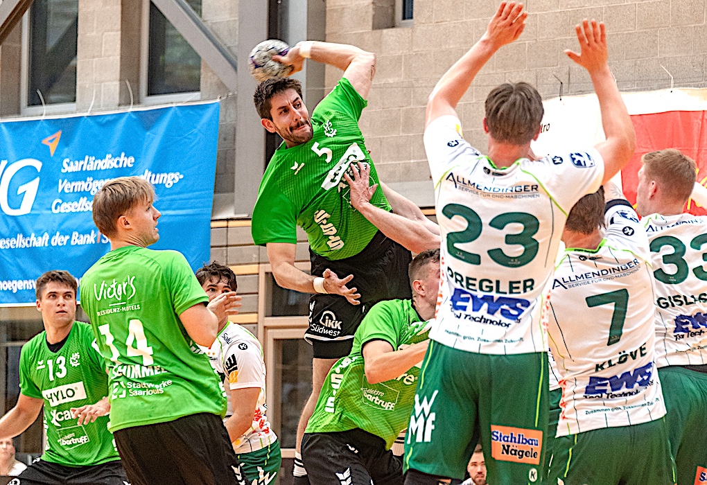 Handballspieler in grünem Trikot setzt zum Sprungwurf an, während mehrere Gegenspieler in weißen Trikots versuchen, ihn zu blocken.