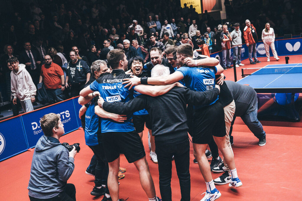 Tischtennisspieler und Betreuer in blauen Trikots liegen sich jubelnd am Spielfeldrand in den Armen, während Zuschauer die Szene verfolgen.