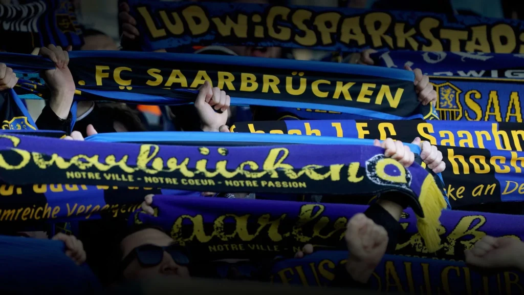 Fußballfans halten blau-gelbe Schals des 1. FC Saarbrücken und mit Saarbrücken-Schriftzügen auf der Tribüne in die Höhe.