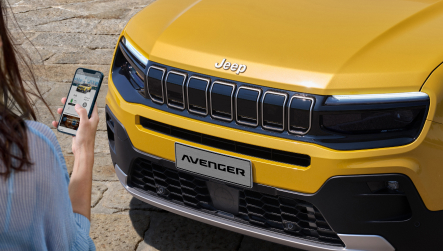 Die Front eines gelben Jeep Avenger Elektro mit markantem siebenteiligem Kühlergrill, schmalen LED-Scheinwerfern und schwarzer Stoßstange. Links im Bild hält eine Person ein Smartphone mit geöffneter App in Richtung des Fahrzeugs.