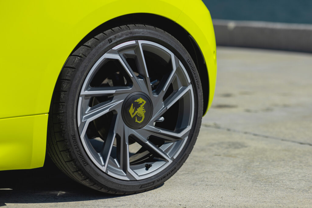 Detailaufnahme eines Rades am gelbgrünen Abarth 500e Turismo. Die mehrspeichige Leichtmetallfelge besitzt ein kantiges Design in Grau und Silber. In der Radmitte befindet sich das gelbe Abarth-Skorpion-Logo, umgeben von einem Bridgestone-Reifen.