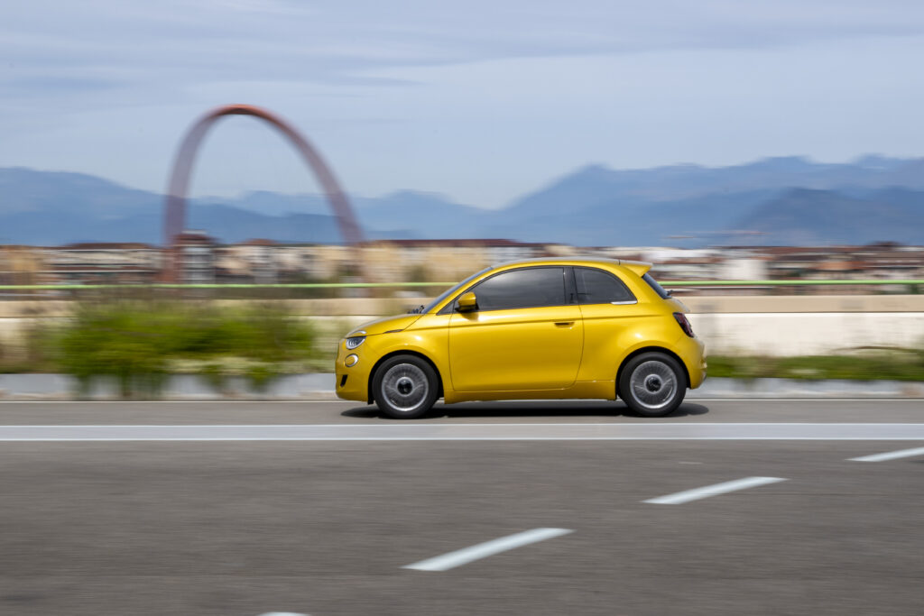 Gelber Fiat 500 fährt auf einer mehrspurigen Straße. Im Hintergrund sind eine Stadtlandschaft, Berge und eine große bogenförmige Konstruktion zu erkennen.