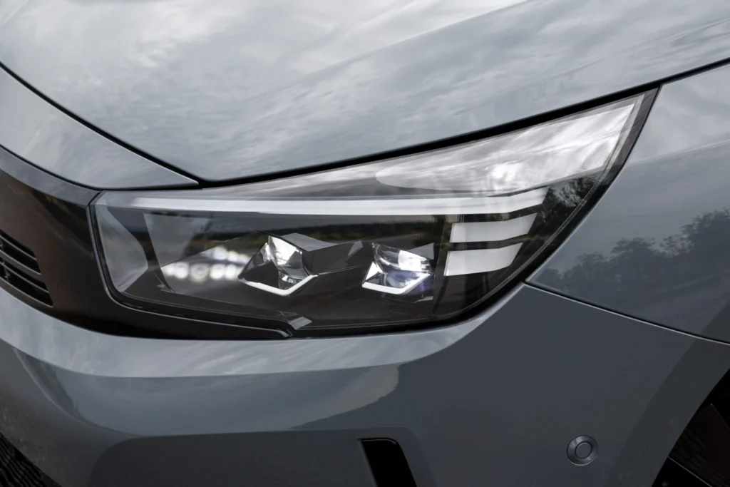 Detailansicht des vorderen Scheinwerfers eines grauen Opel Corsa. Im Gehäuse sind mehrere Leuchtelemente und helle Lichtmodule erkennbar. Der Scheinwerfer ist in die graue Karosserie und die schwarze Frontverkleidung integriert.