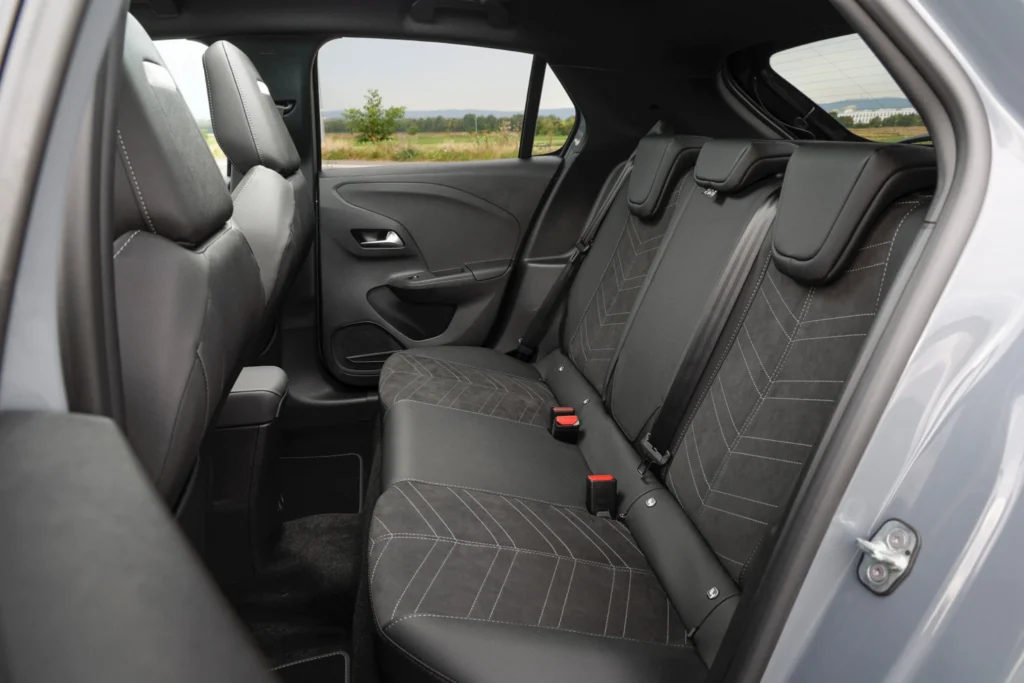 Blick in den Fond eines Opel Corsa mit dreisitziger Rückbank und dunkler Stoffpolsterung mit hellen Ziernähten. Drei Kopfstützen, Sicherheitsgurte und rote Gurtschlösser sind sichtbar. Durch die Fenster ist eine grüne Landschaft zu erkennen.