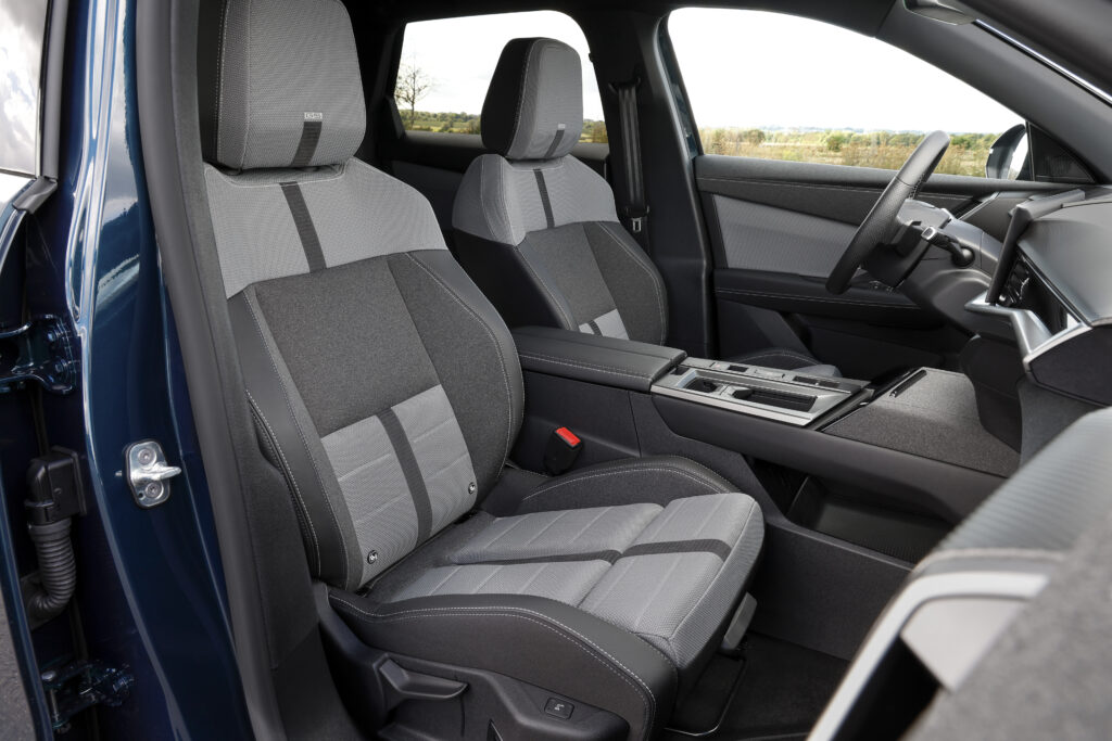 Blick durch die geöffnete Seitentür auf die beiden Vordersitze des Opel Grandland Hybrid. Die Sitze sind mit grau-schwarzem Stoff bezogen und verfügen über kontrastierende Ziernähte. Daneben sind die Mittelkonsole und das Lenkrad zu erkennen.