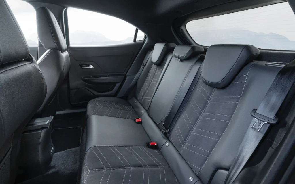 Blick auf die dreisitzige Rückbank des Opel Mokka Electric mit dunkler Polsterung, hellen Ziernähten und integrierten Kopfstützen. Drei Sicherheitsgurte und rote Gurtschlösser sind sichtbar. Durch die Fenster ist eine helle Berglandschaft zu erkennen.