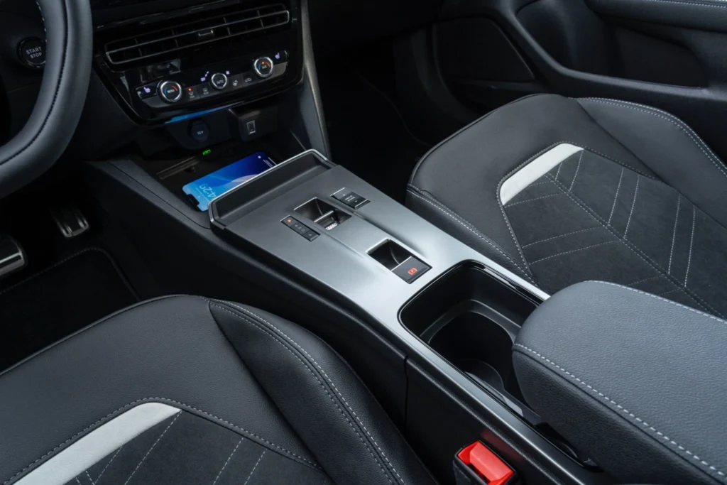 Detailansicht der Mittelkonsole im Opel Mokka Electric mit silberfarbener Oberfläche, elektronischem Gangwahlschalter, elektrischer Parkbremse und zwei Getränkehaltern. Im vorderen Ablagefach liegt ein Smartphone; daneben sind die dunkel gemusterten Vordersitze zu sehen.